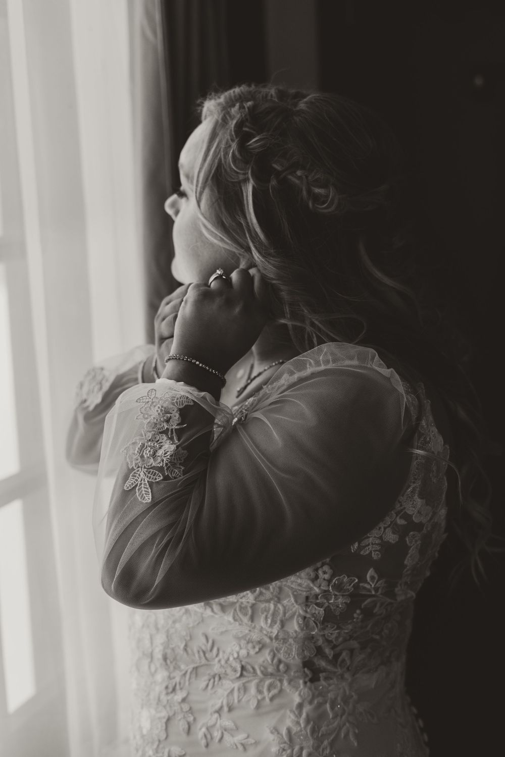 Black and white portrait of bride in lace dress adjusting earring by window light.