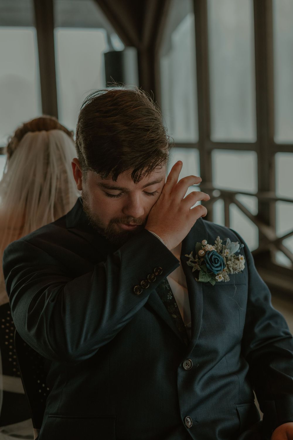 Emotional groom in dark suit with floral boutonniere wiping tears during wedding ceremony.