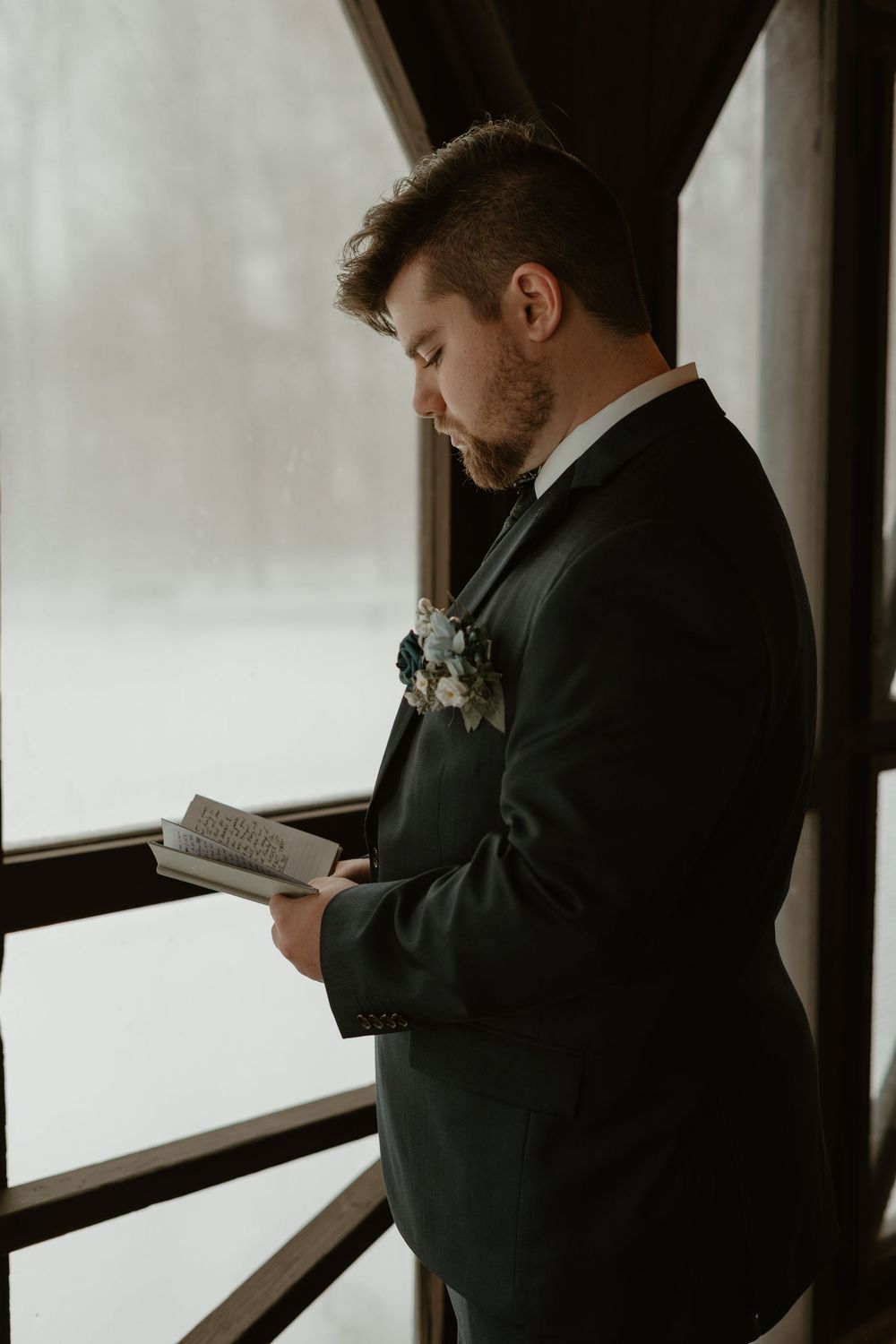 Groom in dark suit reading vows by window with blue floral boutonniere in snowy setting.