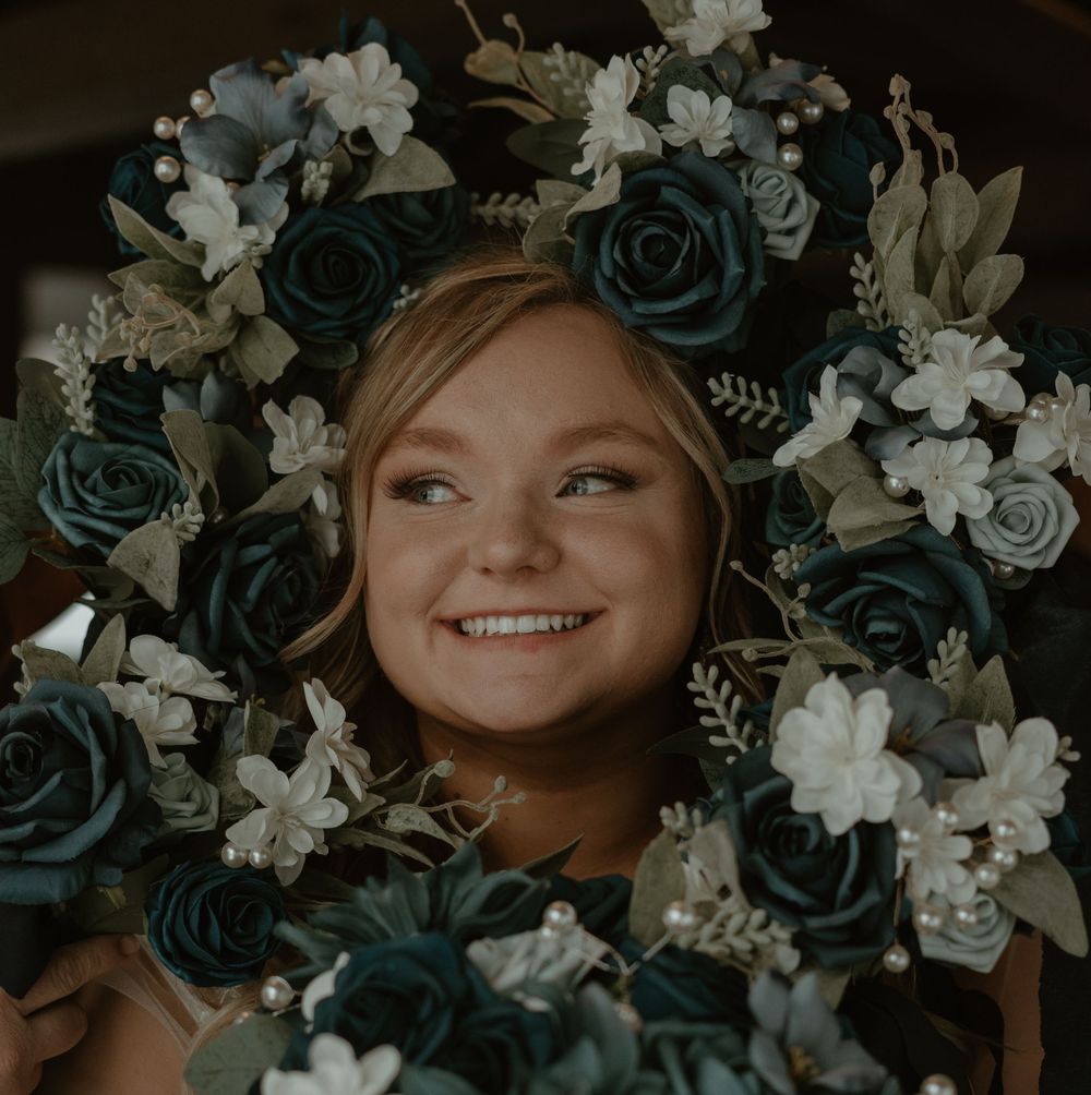 Smiling bridesmaid surrounded by teal and white floral wedding wreaths in joyful portrait.