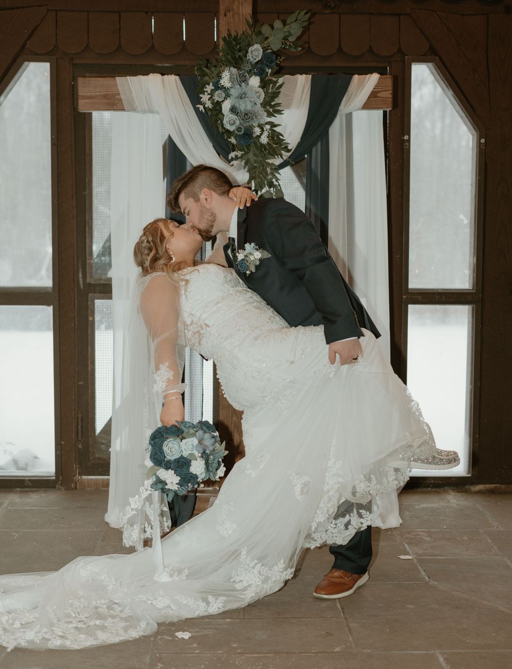 Bride and groom sharing romantic dip kiss at altar decorated with blue floral arch.