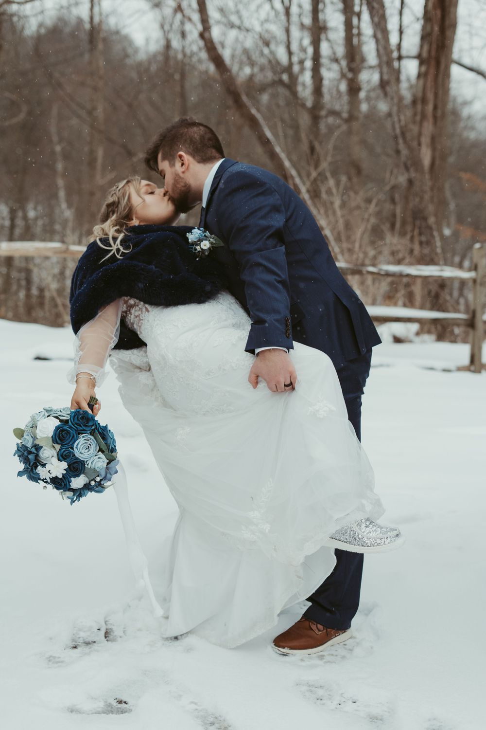 Bride and groom kissing in snowy winter landscape, bride holding blue and white bouquet.