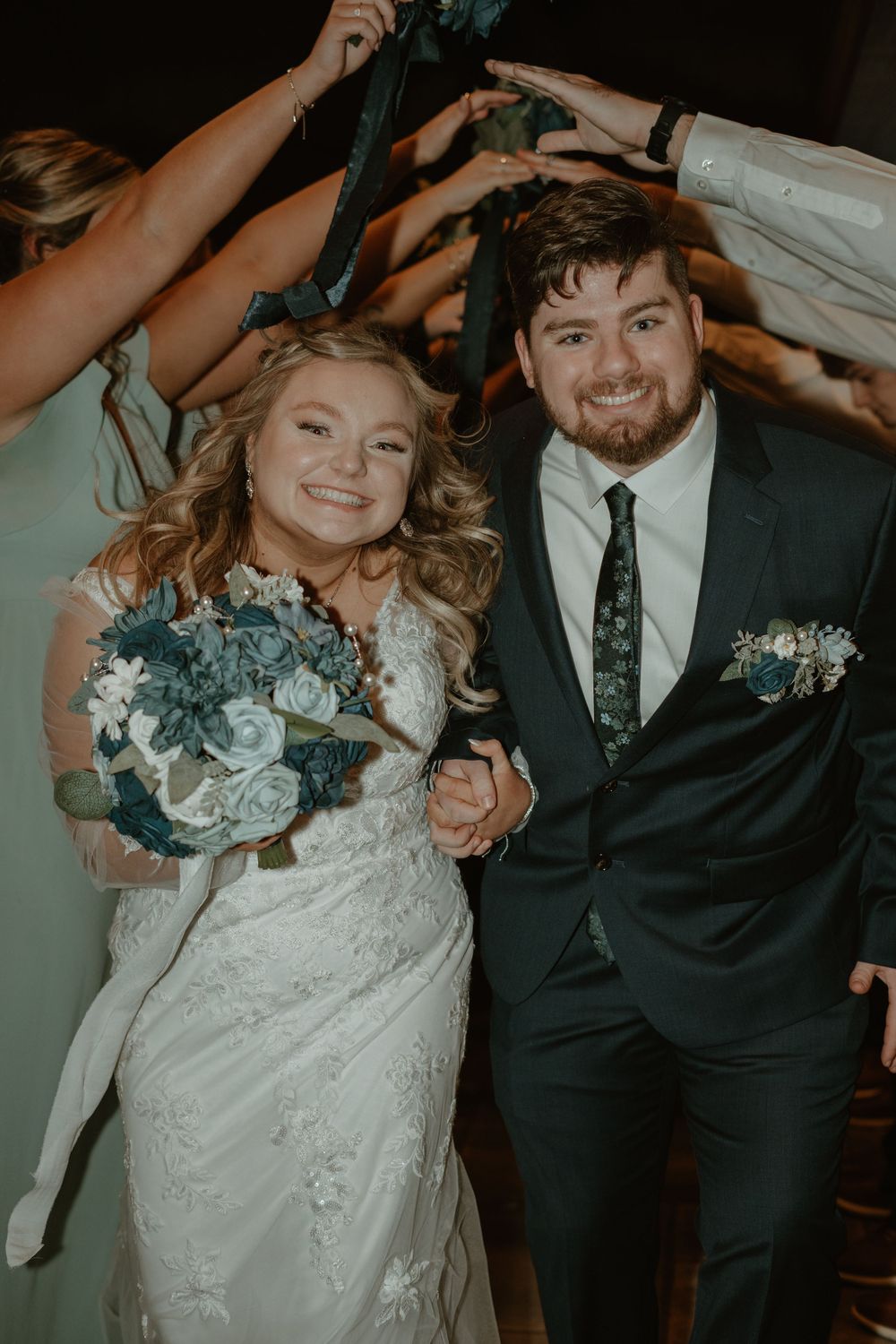 Smiling bride and groom walk through tunnel of raised hands at wedding reception, bride holding teal floral bouquet.