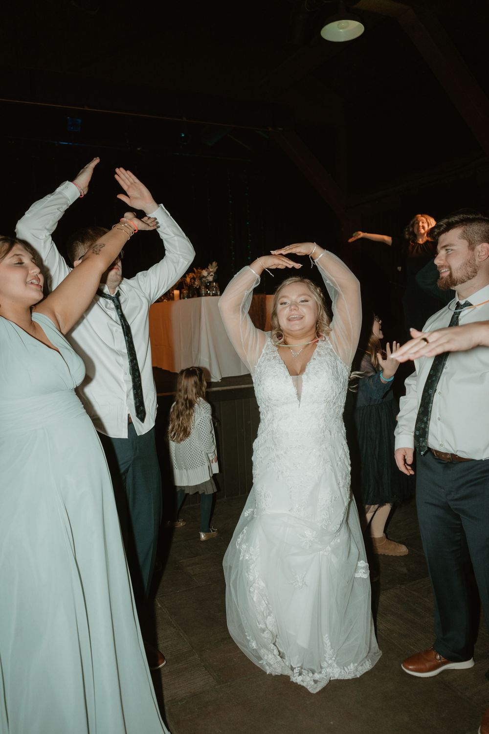 Bride in lace wedding gown dances with arms raised at wedding reception surrounded by guests.