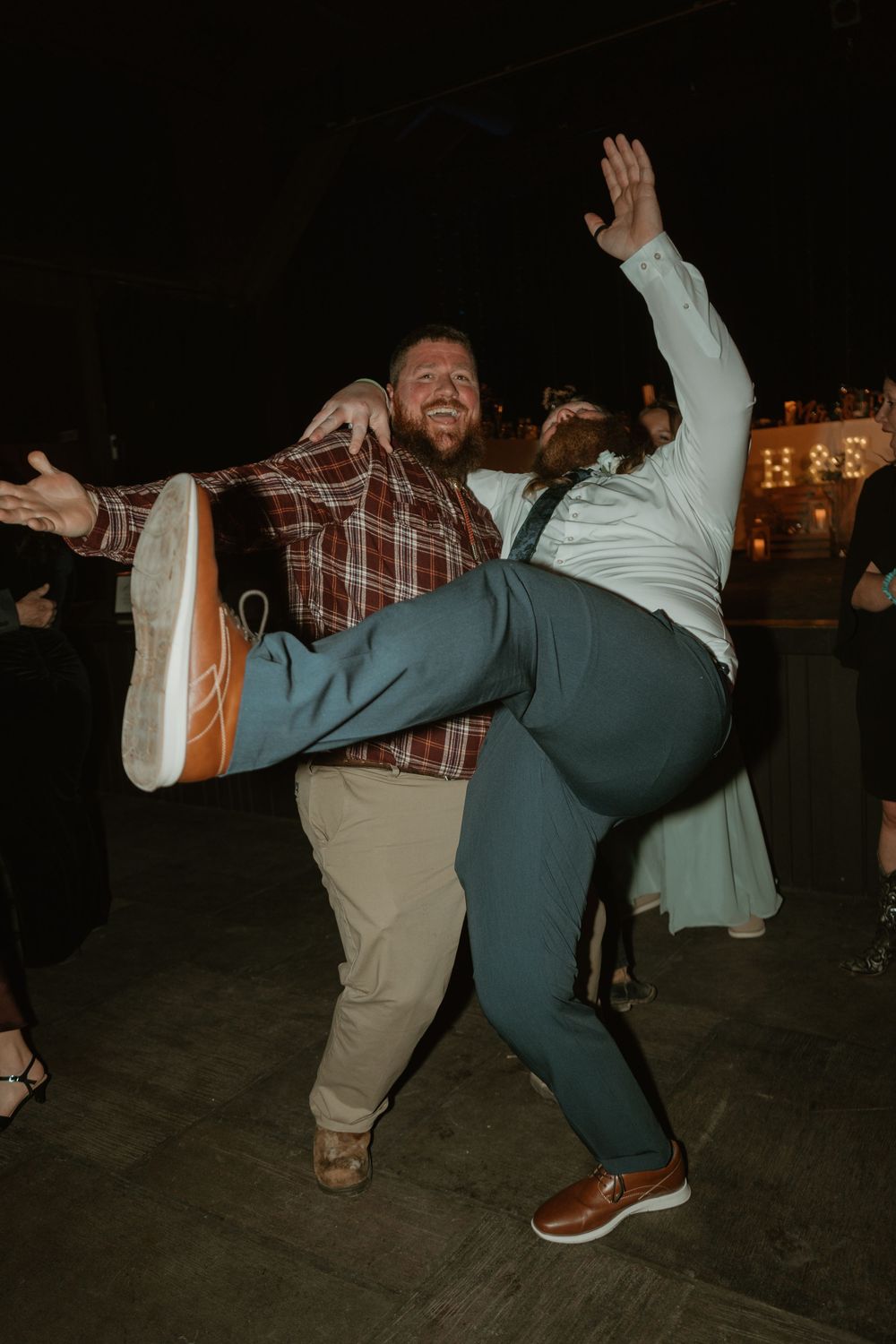 Two men energetically dancing at outdoor wedding reception at night, one lifting the other.