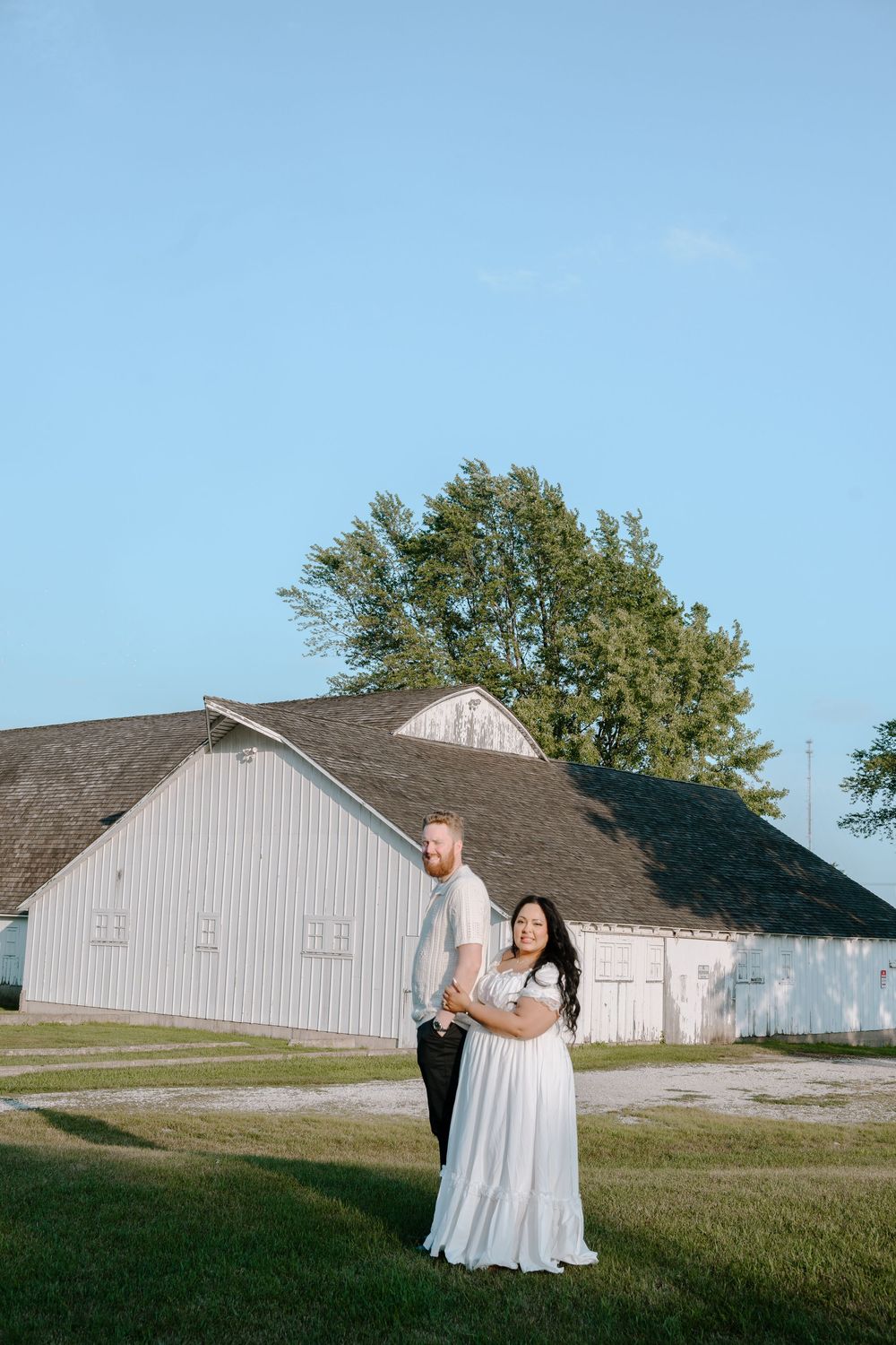 A couple shares an intimate dance together on a grassy field with white farm buildings in the background during sunset.