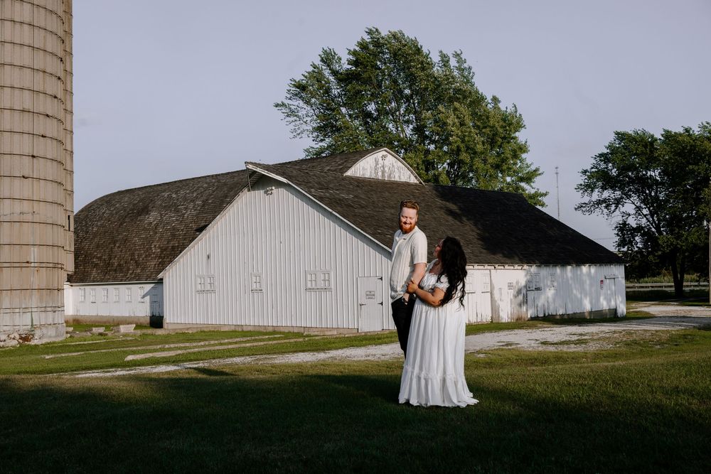 A romantic couple poses in front of a rustic white barn and silo during a countryside photoshoot.