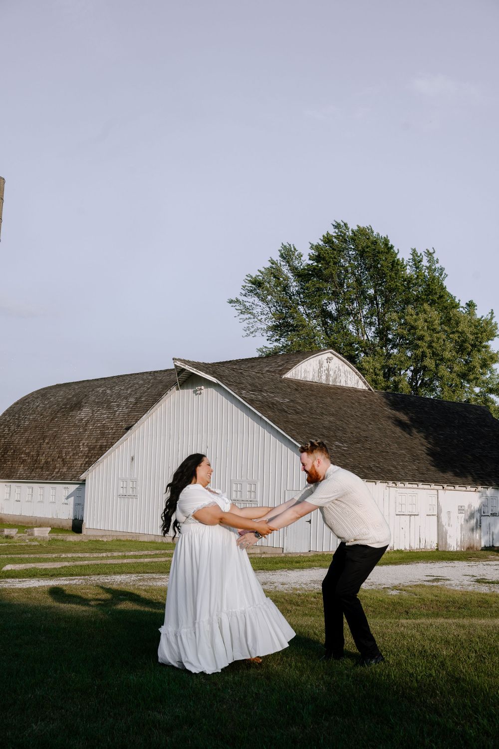 A playful couple in white and black attire pose dramatically in front of a rustic barn on a grassy field.