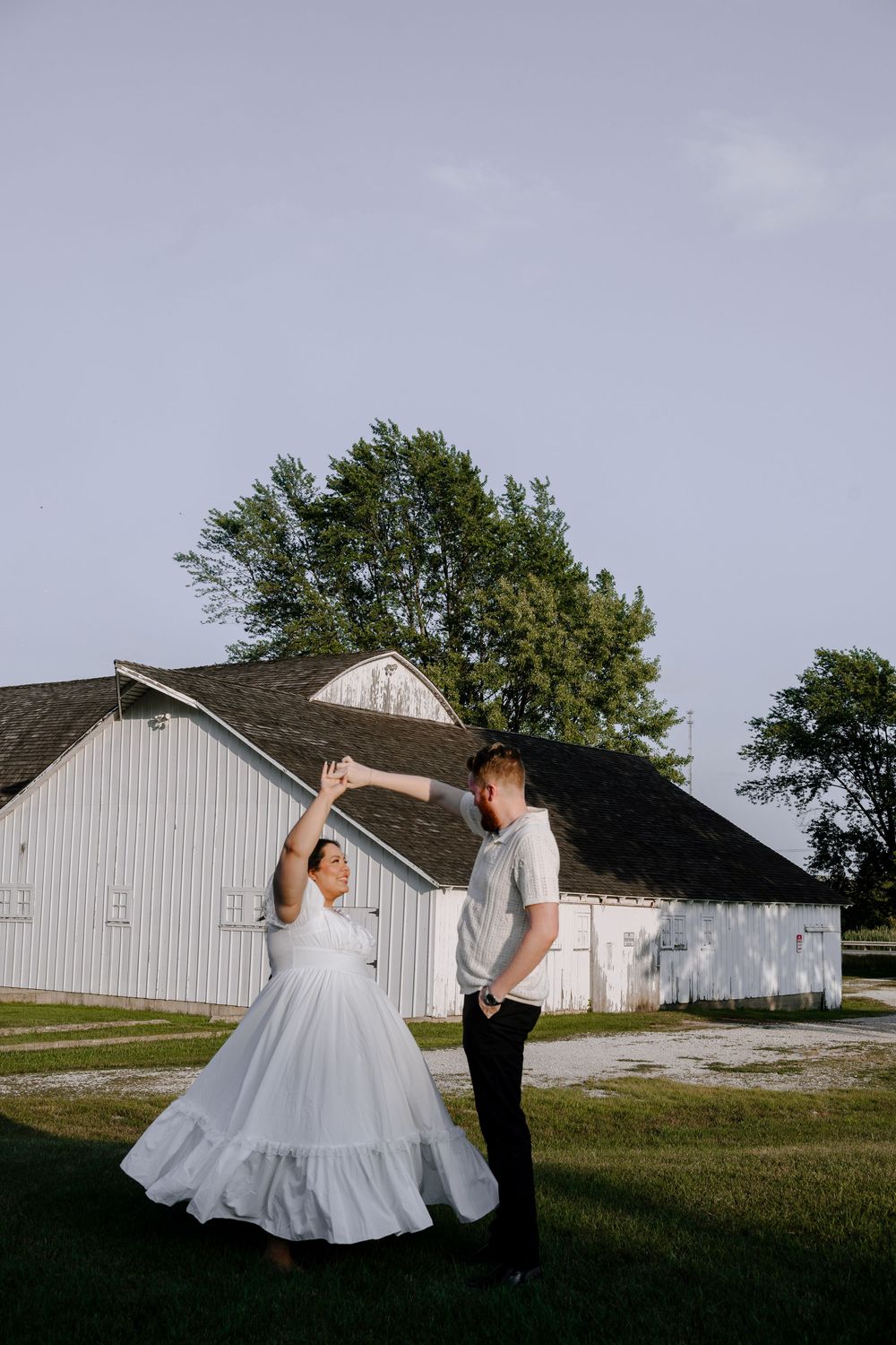 A couple twirls together on grass with white farm buildings in the background.