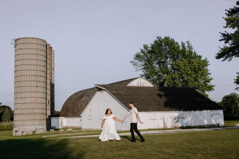 A romantic couple poses near a rustic barn and silo on a grassy hill during a countryside photo session.
