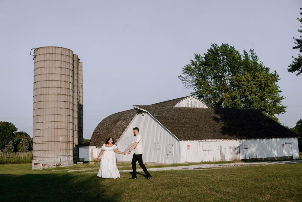 A couple holding hands walks together near a white barn and tall silo in a rural farm setting.