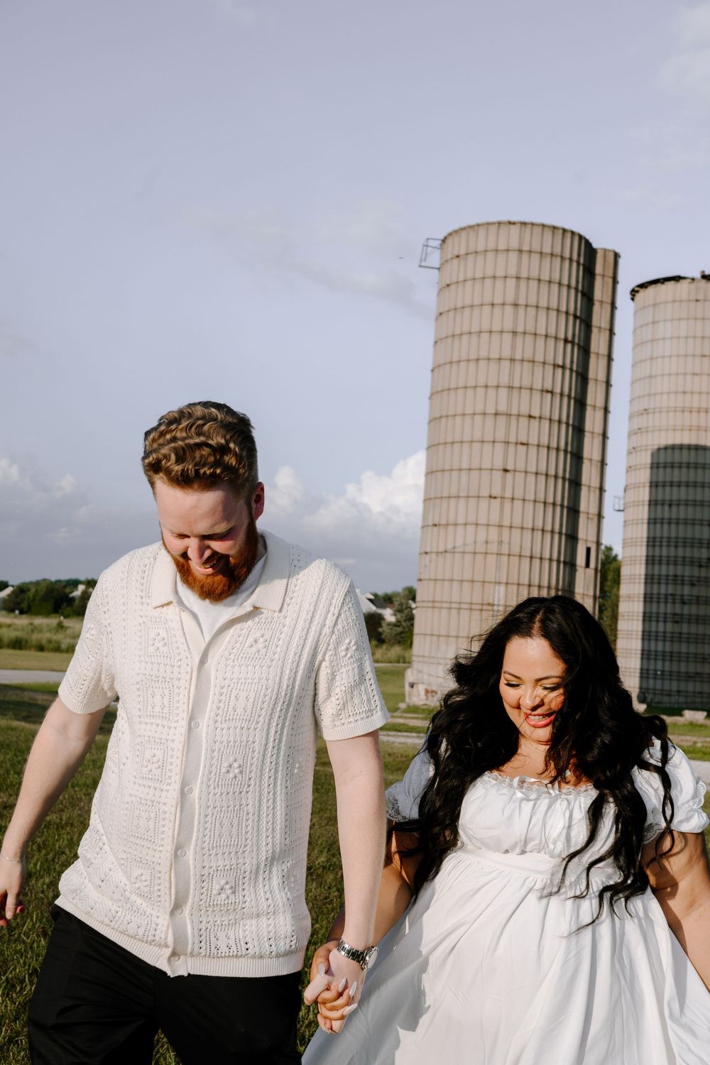 Joyful couple walking hand in hand beside tall farm silos on a cloudy day.