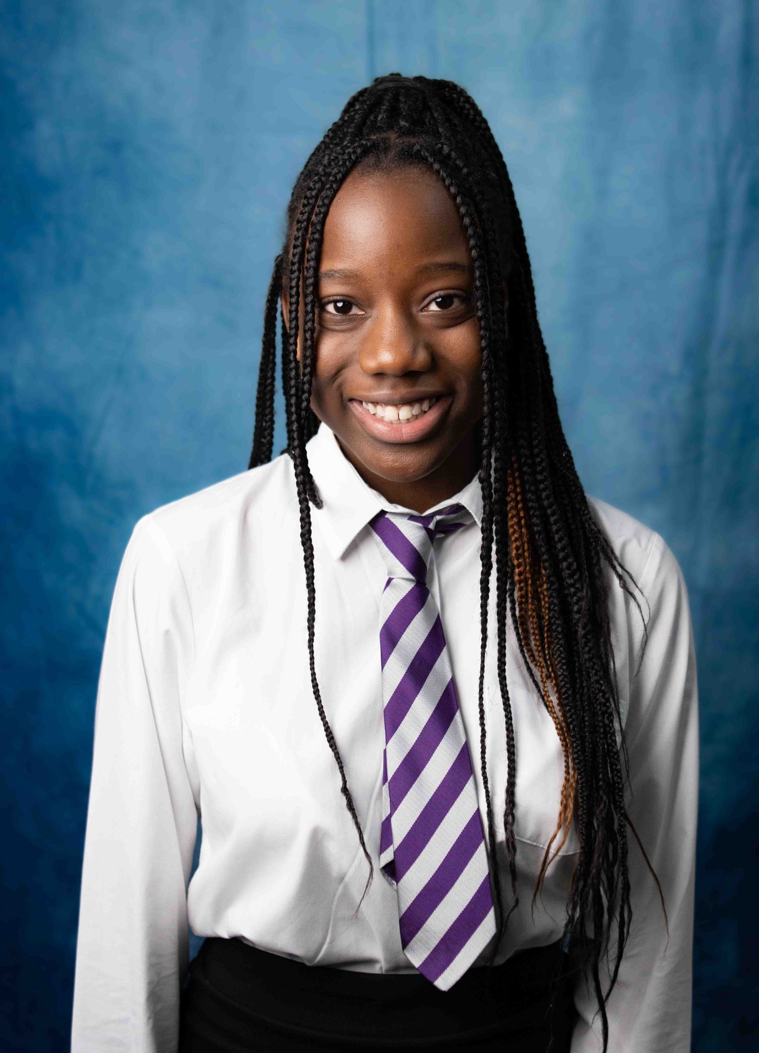 Professional portrait of someone in a white dress shirt and purple striped tie with long braided hair against a blue backdrop.