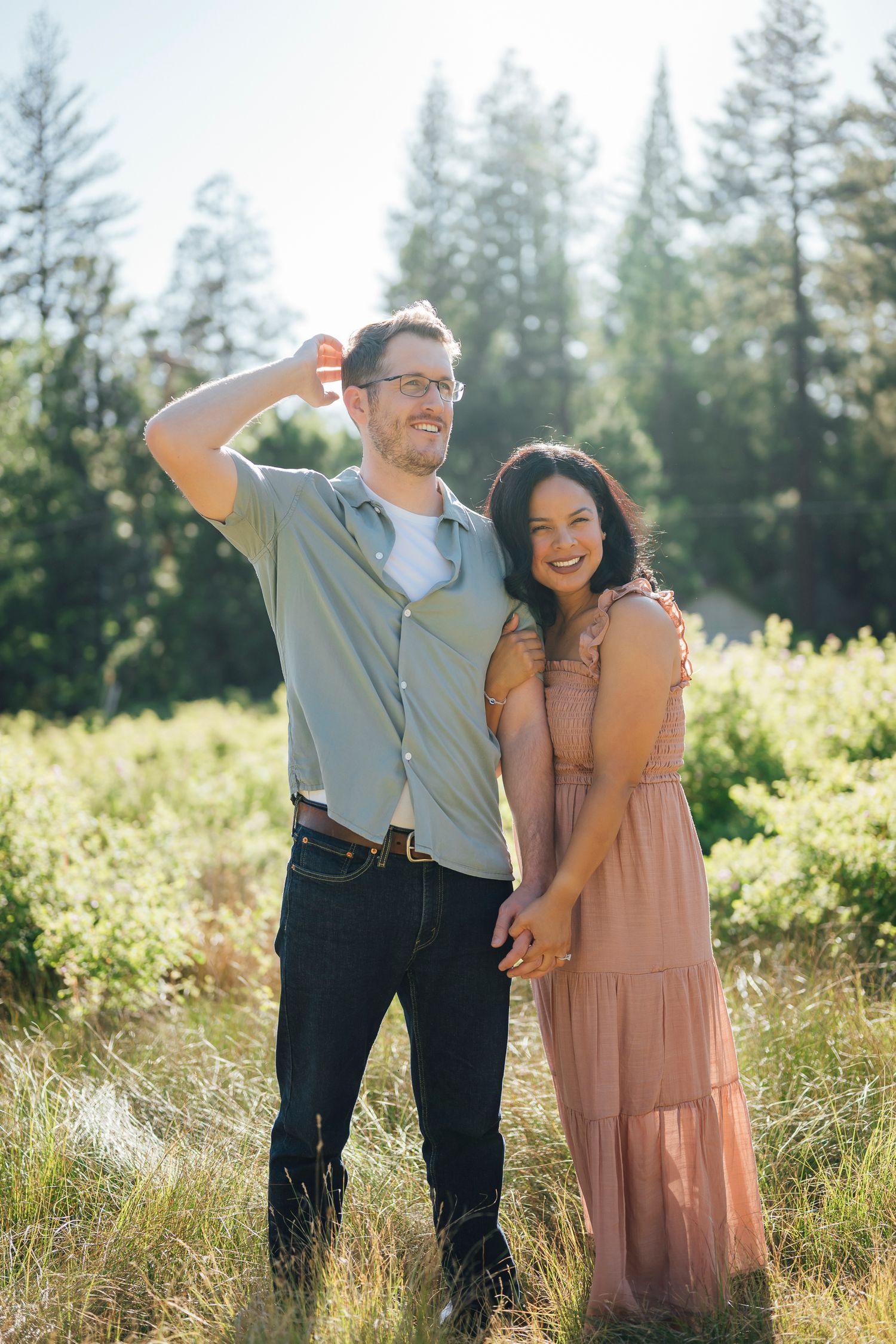 A couple stands together in a sunny meadow with evergreen trees in the background wearing pink and gray outfits.