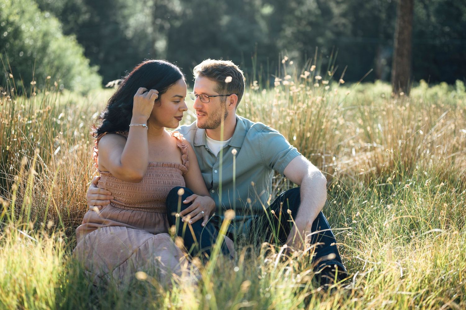 A romantic couple sits close together in a sunlit meadow with tall grass and trees in the background.