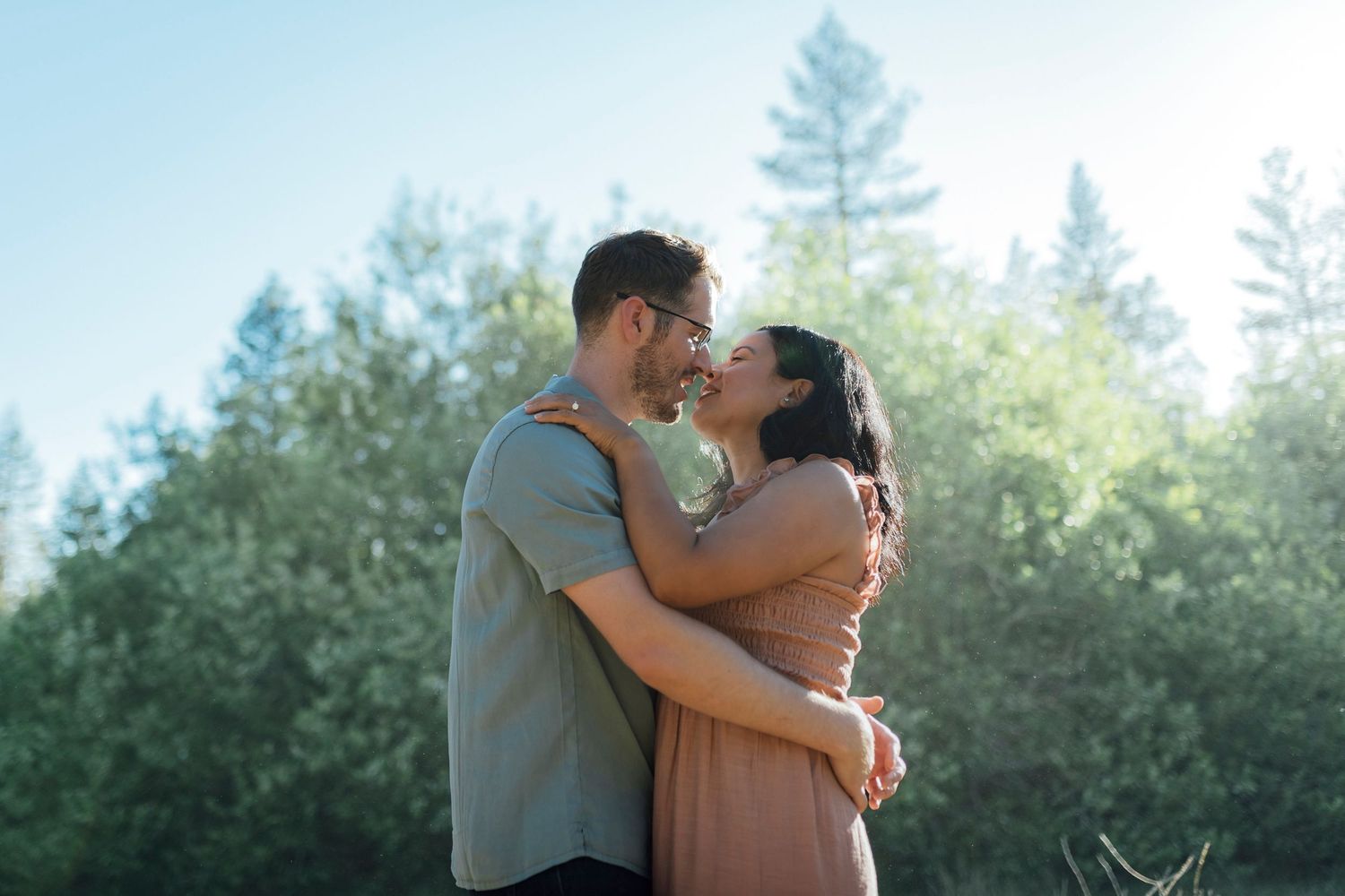A romantic couple embraces outdoors against a backdrop of evergreen trees in soft sunlight.