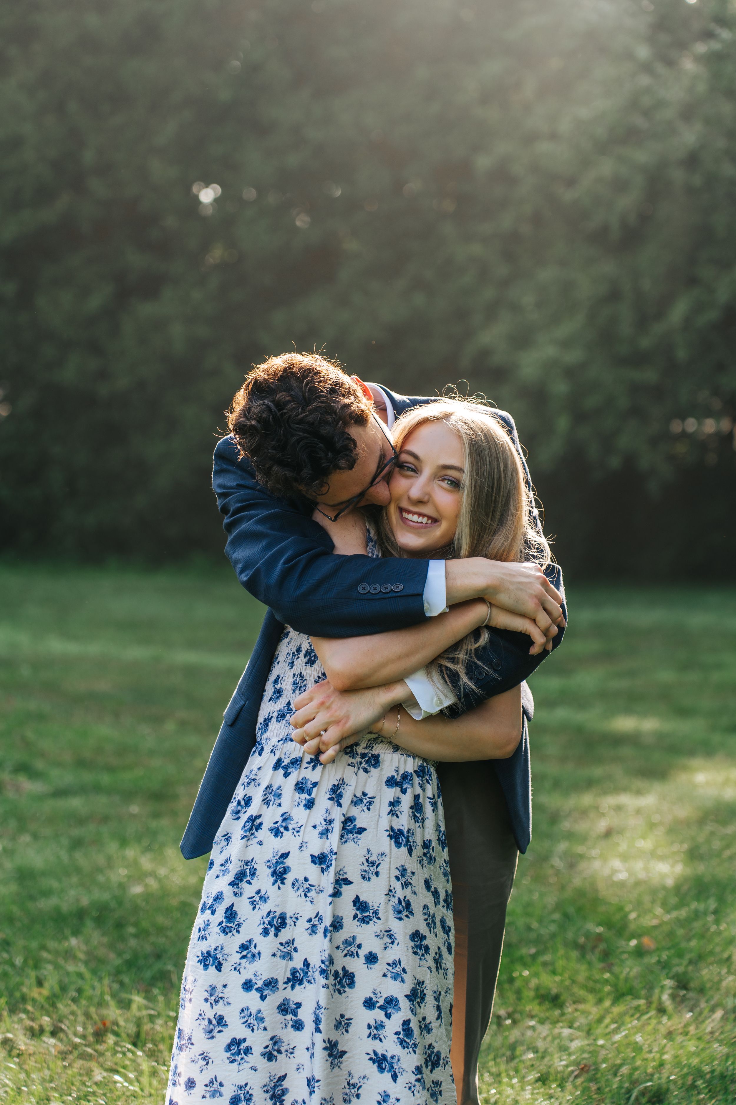 A couple embraces in a sunlit meadow, with the woman wearing a blue and white floral dress and her partner in navy.
