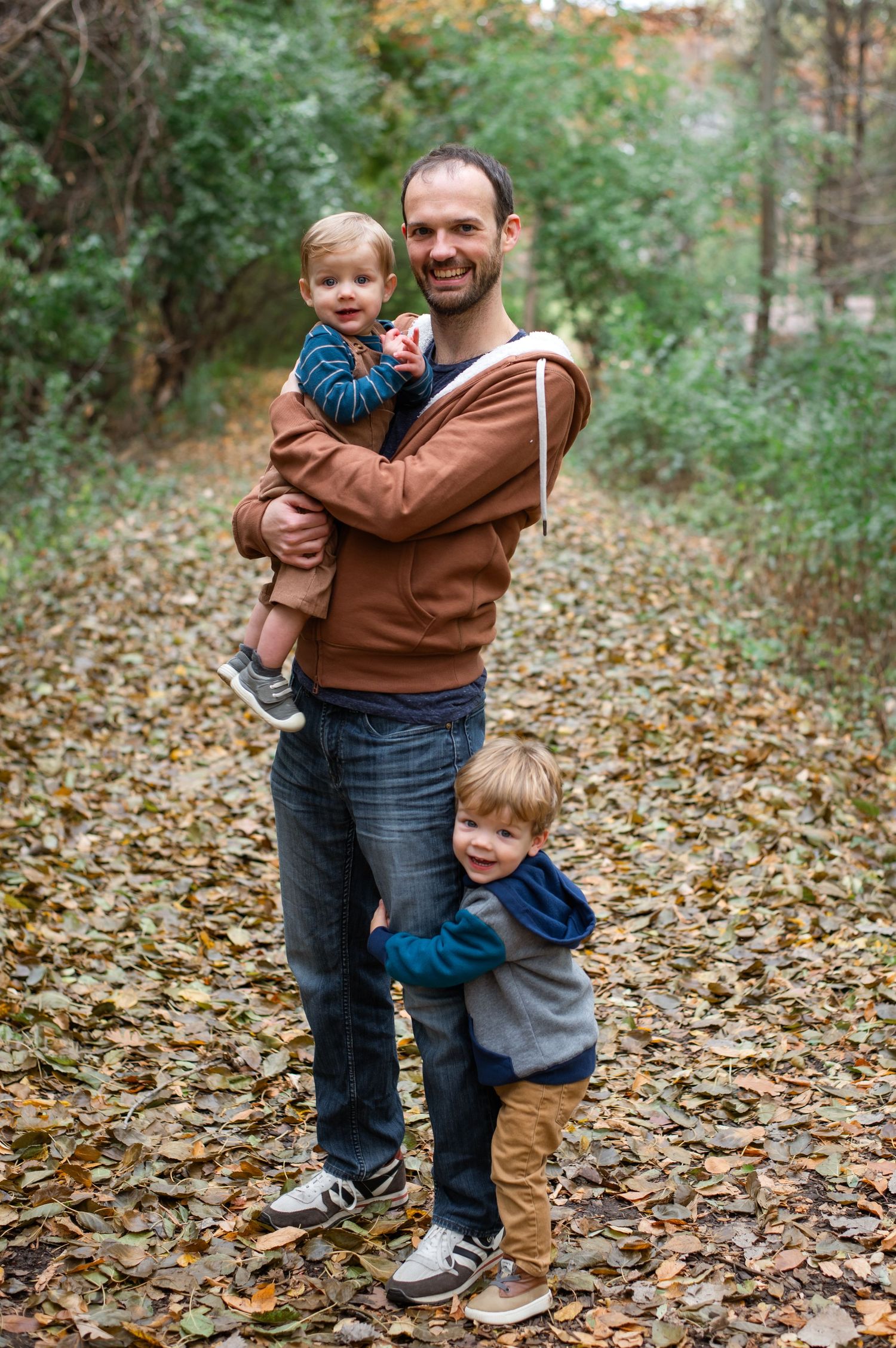 A tender autumn moment on a leaf-covered forest trail shows adults and children sharing embrace and laughter.