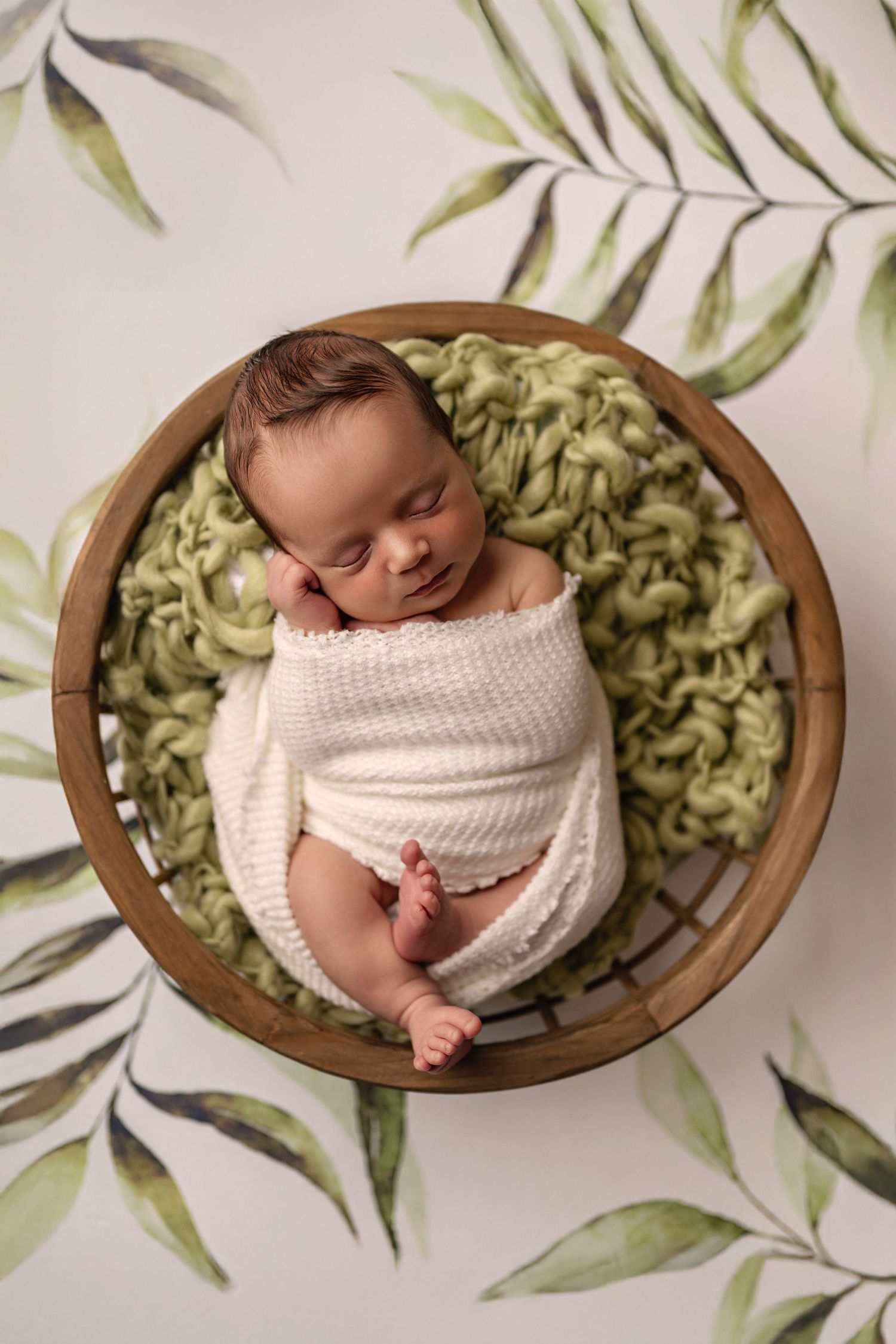 Newborn baby sleeps peacefully in wooden bowl with green blanket and botanical backdrop.