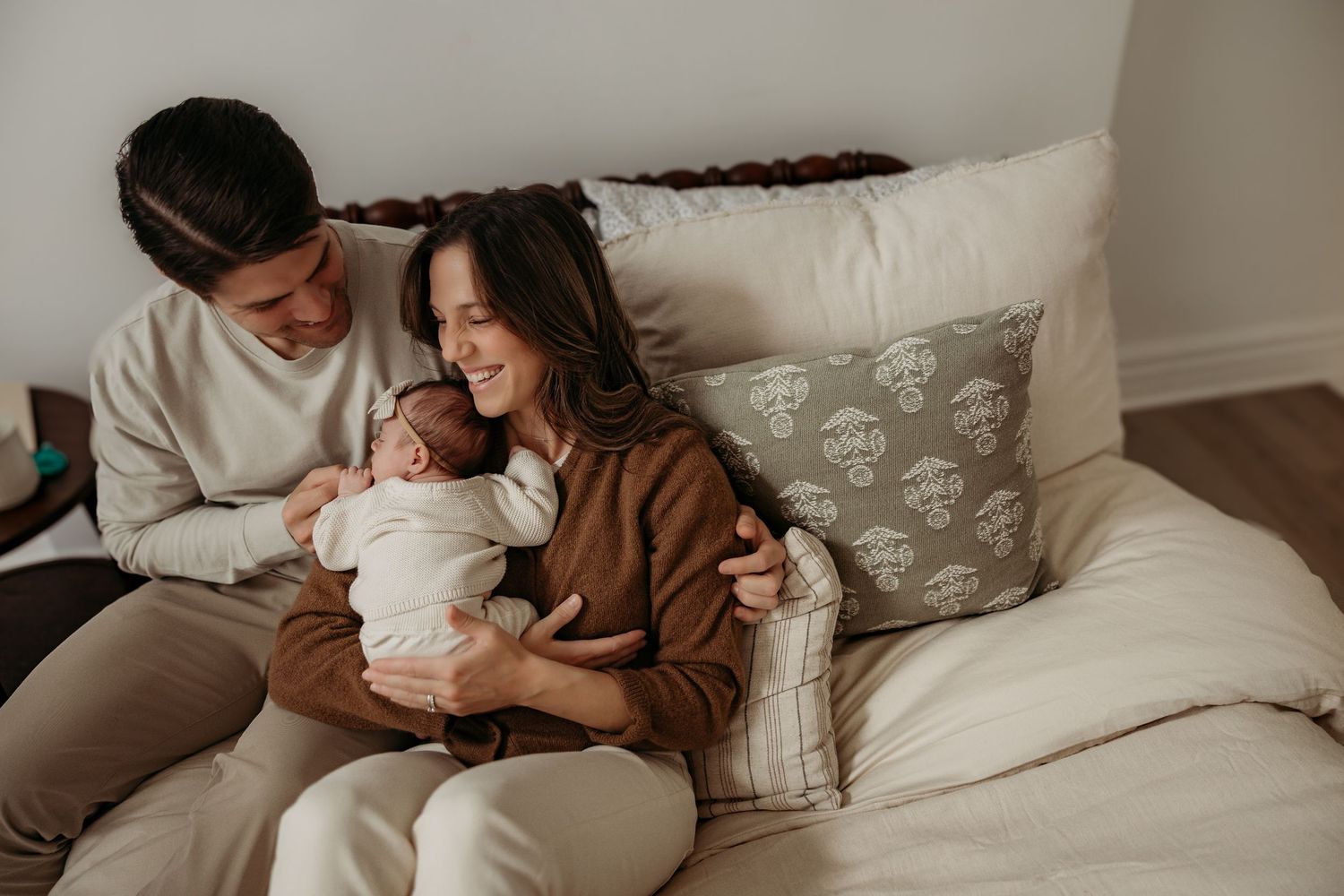 A couple sits on a couch together, holding a sleeping newborn baby while sharing a tender moment at home.