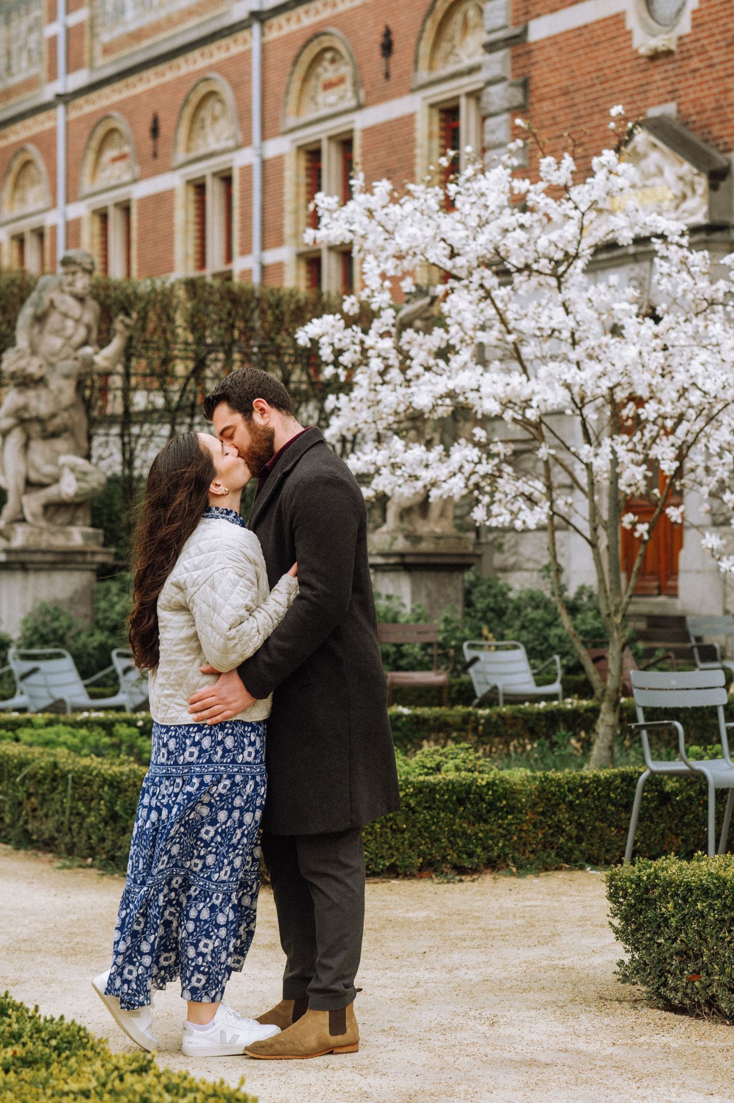 Couple kissing after a secret proposal in the Rijksmuseum gardens with historic statues in the background, Amsterdam.