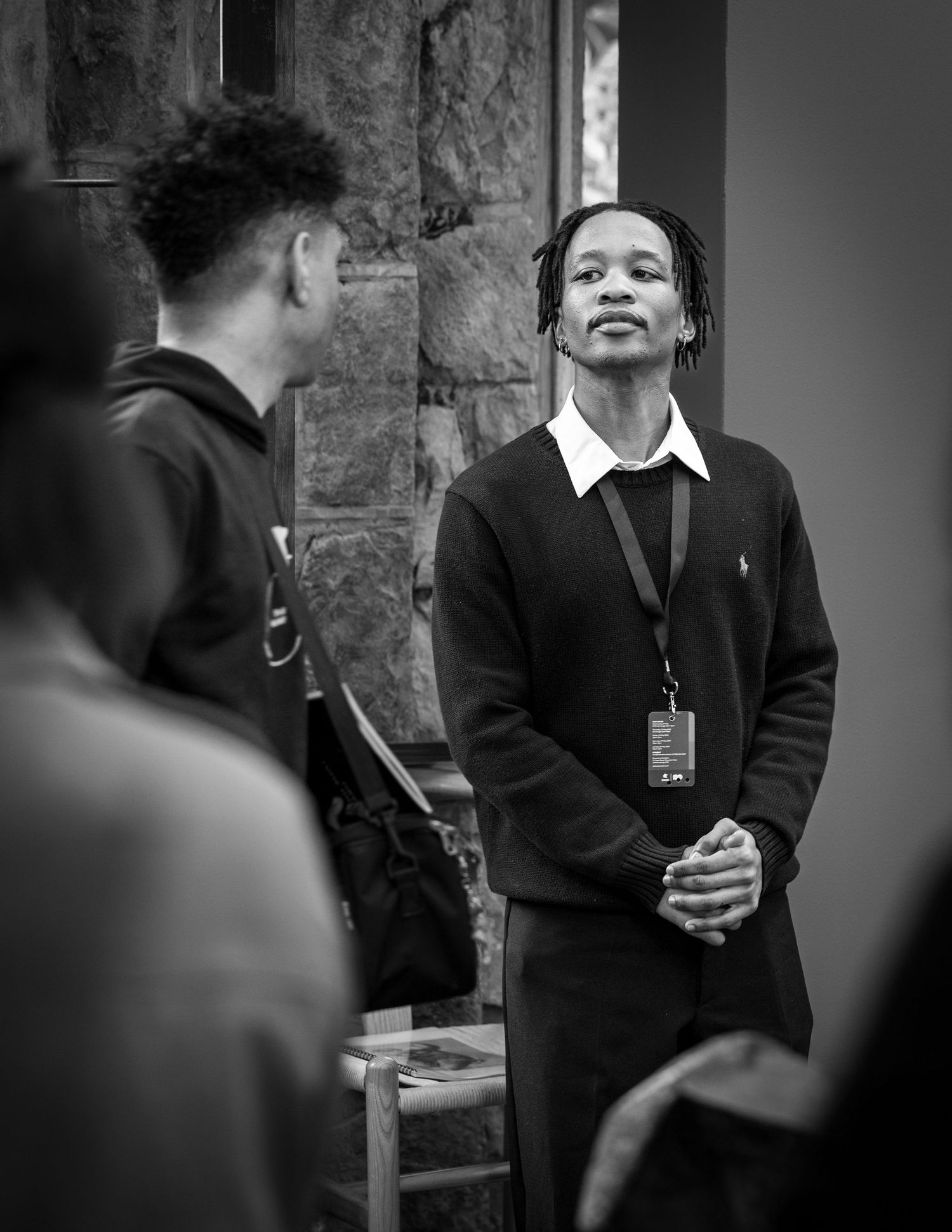 Black and white photograph of two people interacting near a brick wall, one wearing a dark sweater with a lanyard.