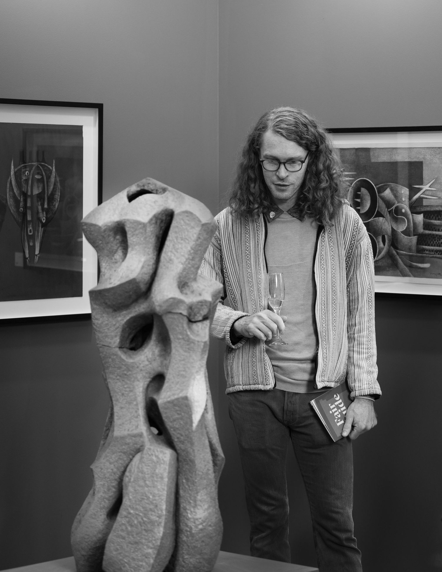 Black and white photo showing a figure standing near an abstract sculptural form and framed artwork in a gallery space.