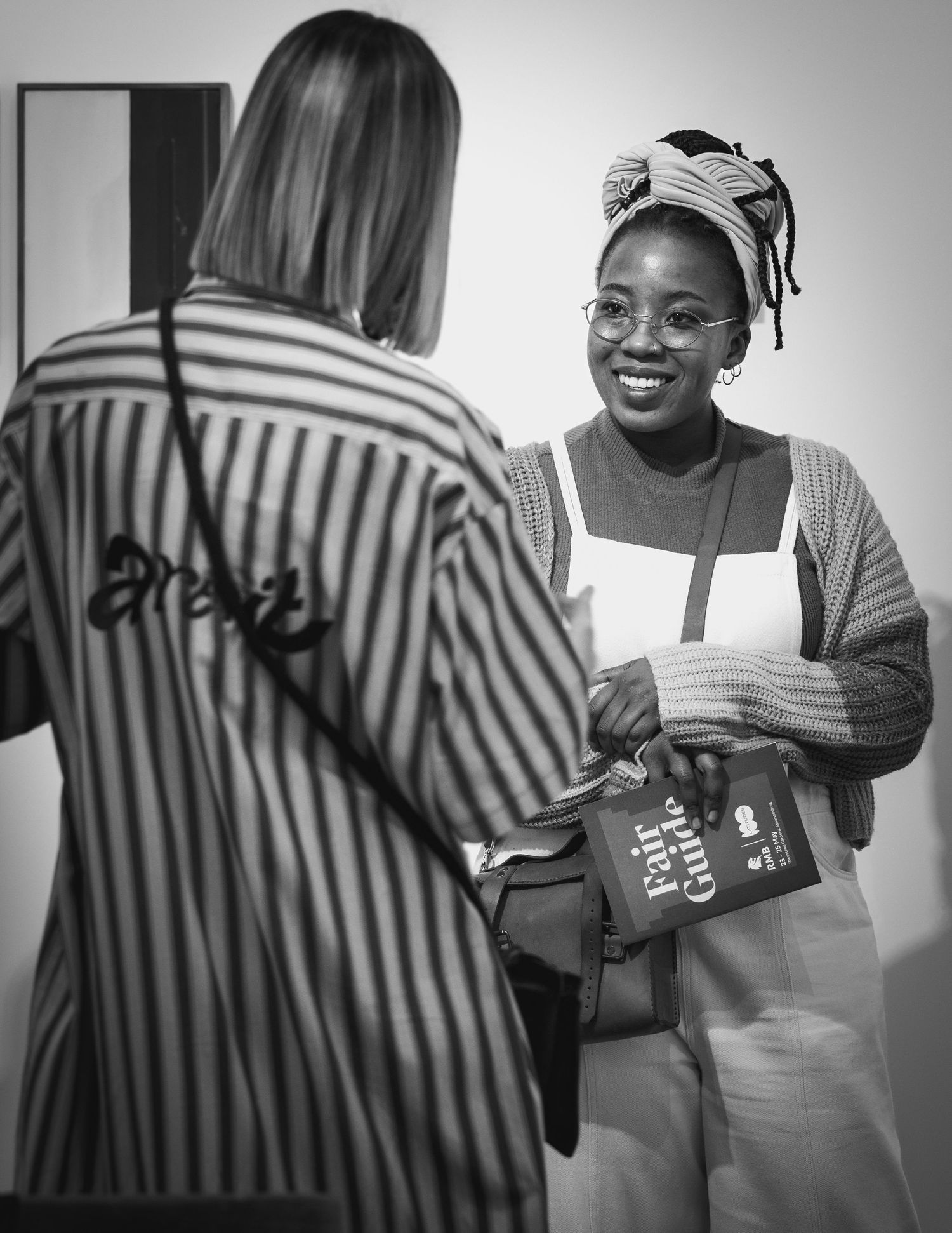 A black and white candid photo of two people in conversation, one wearing a headwrap and smiling while holding papers.