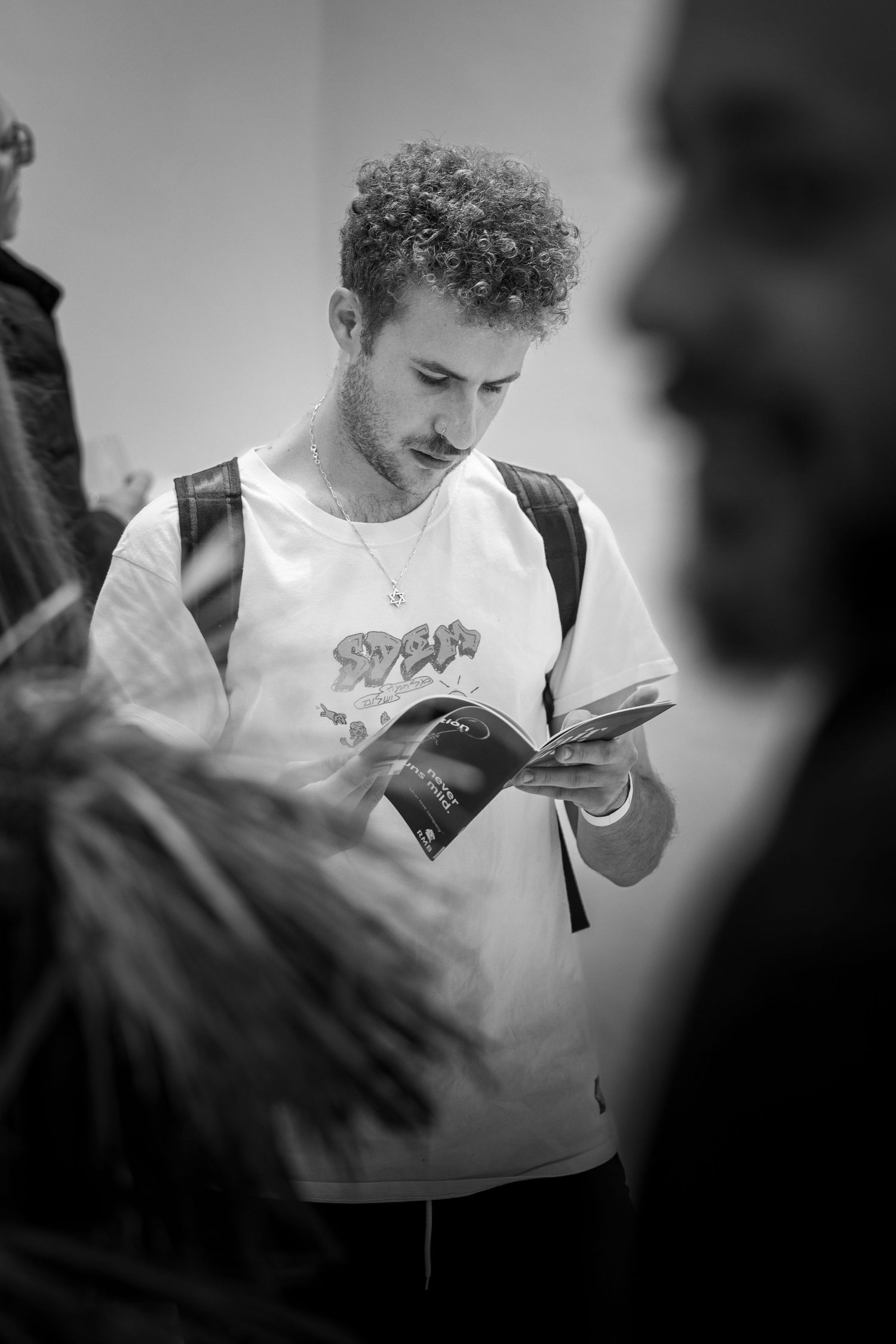 Black and white portrait of a person with curly hair reading a book while wearing a backpack and graphic t-shirt.