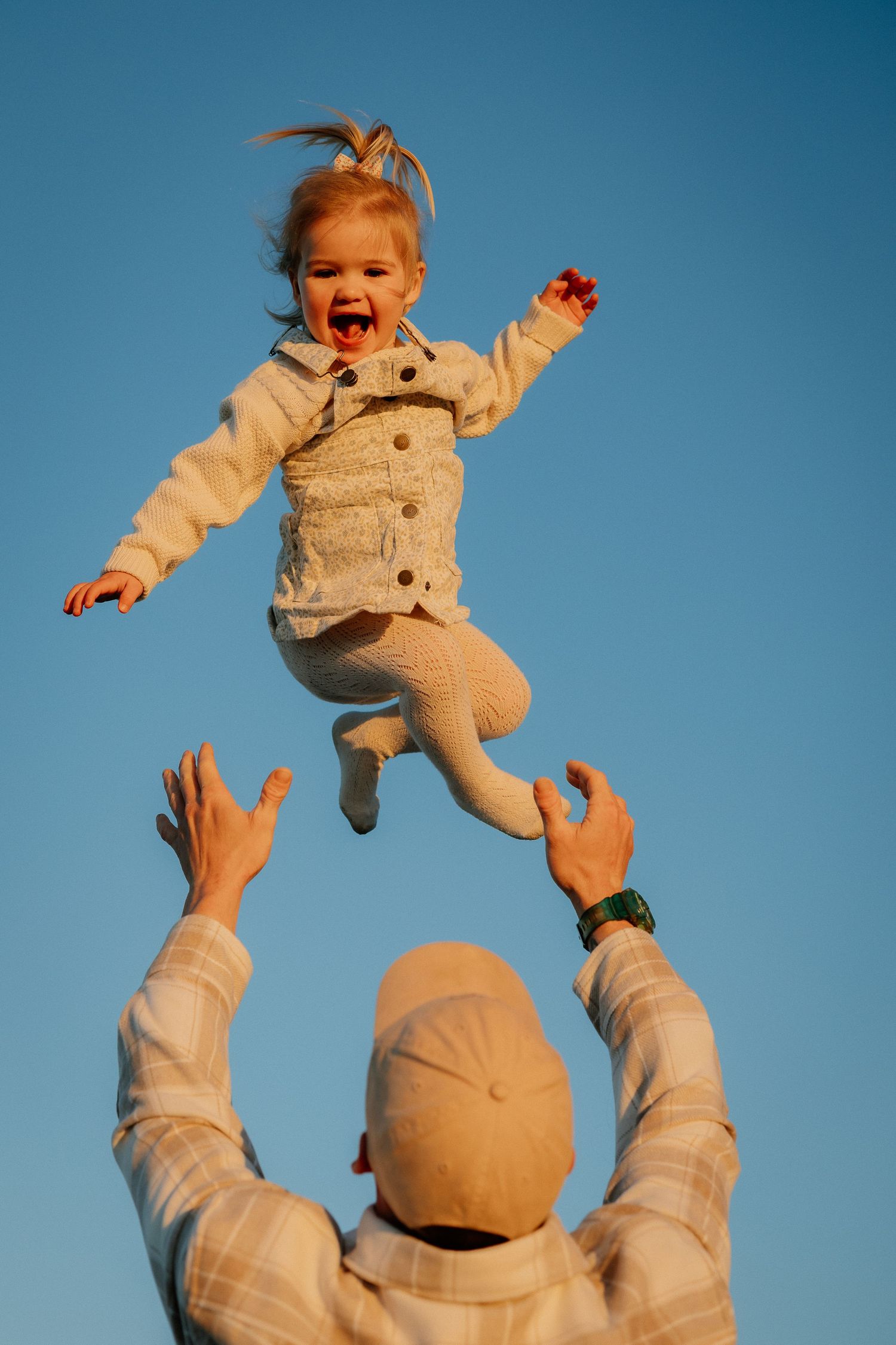 Against a blue sky background, someone tosses a small child wearing beige clothing joyfully into the air.