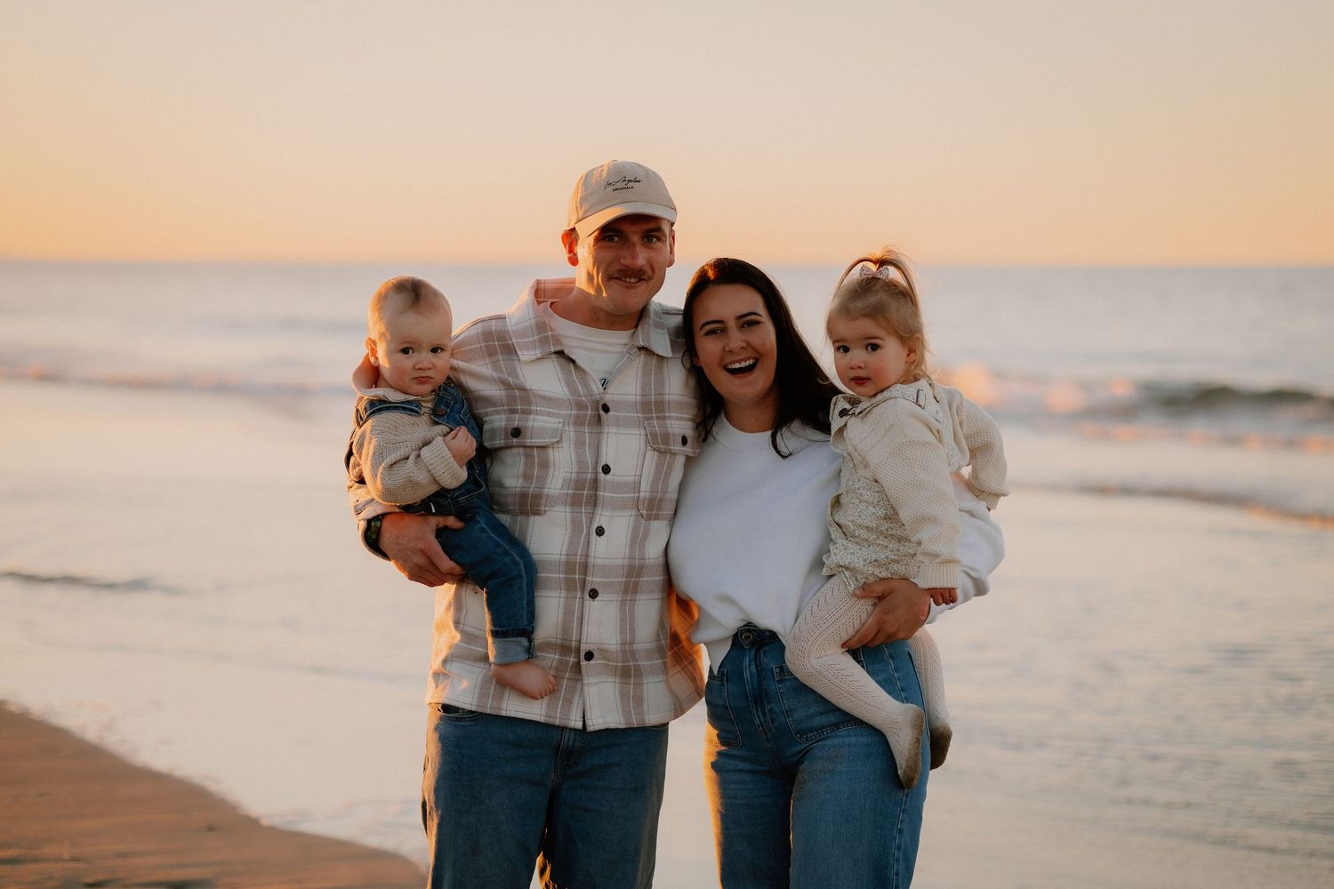 Family wearing coordinated blue and neutral outfits walking together on beach at sunset.