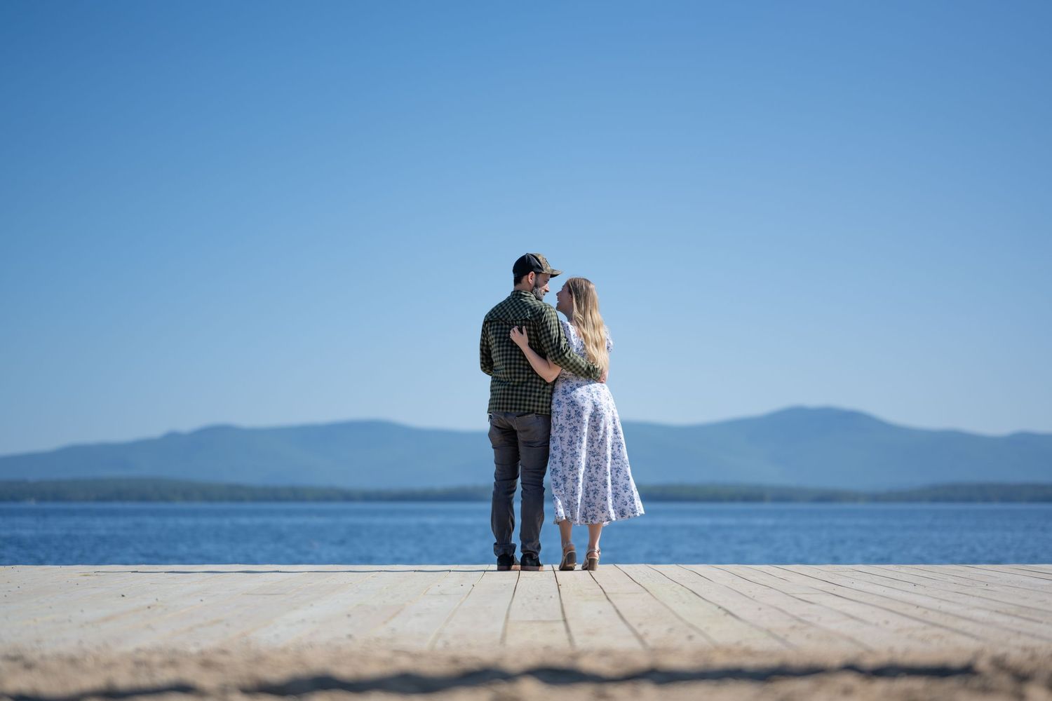 Romantic couple embracing on a wooden dock overlooking a serene lake with mountain views.