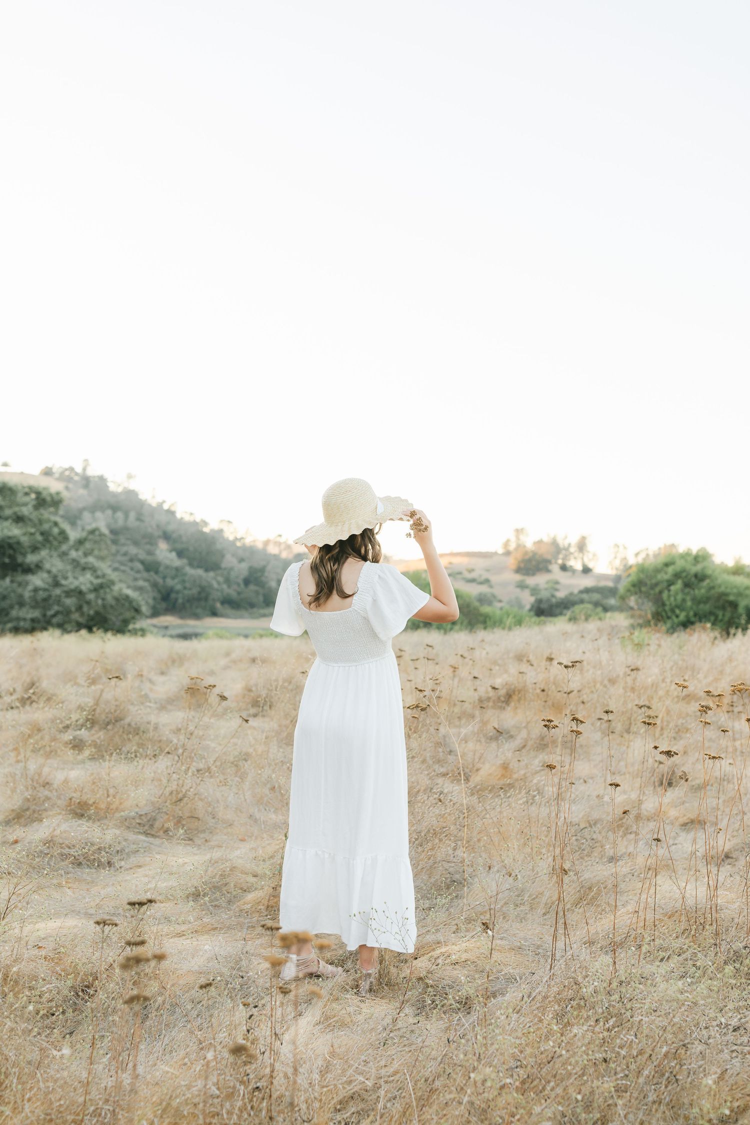Woman in white dress stands thoughtfully in golden field of dry grass during sunset, creating ethereal summer scene captured by Chelsee Raw.