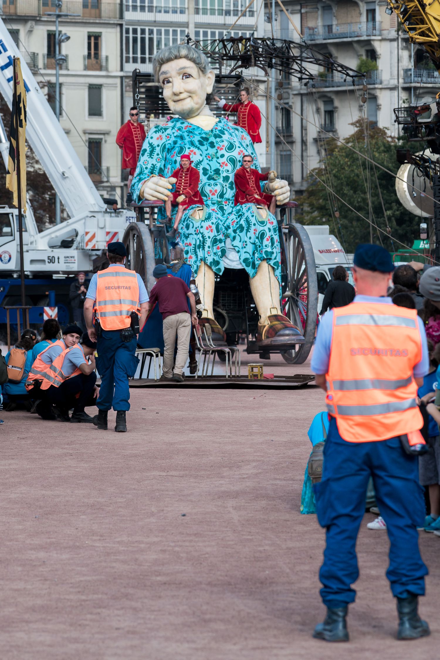 Workers in orange safety vests and blue uniforms walk in a public square with buildings in the background.