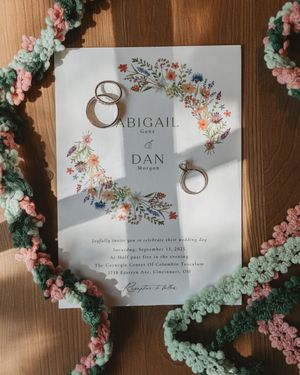 Wedding invitation cards with floral designs and wedding rings displayed on wooden surface surrounded by delicate flowers.