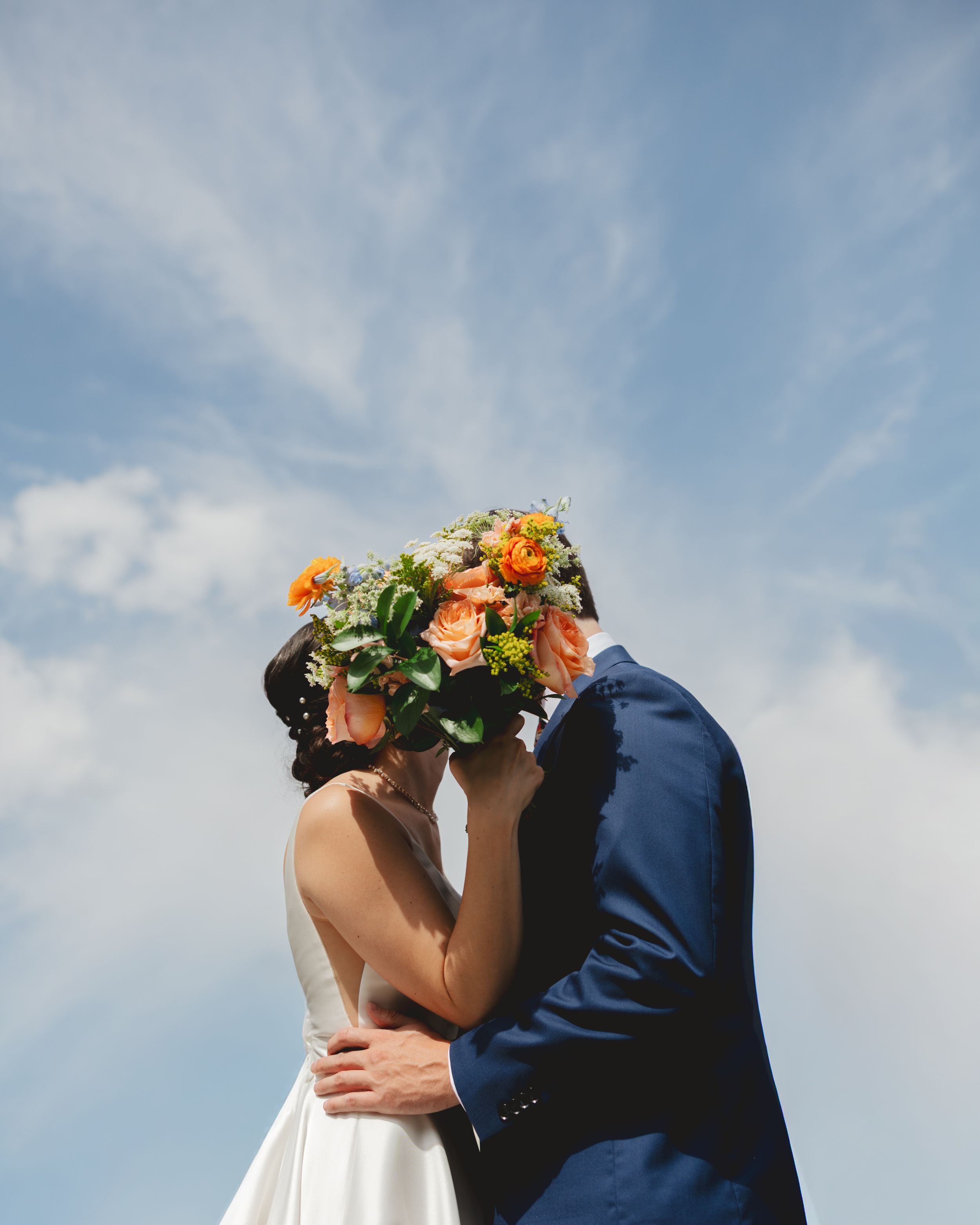 A romantic wedding couple shares a tender kiss against a soft blue sky backdrop with wispy clouds.