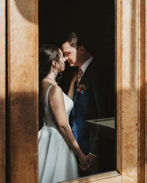 Romantic wedding couple stands in soft window light casting shadows on wooden paneled walls.