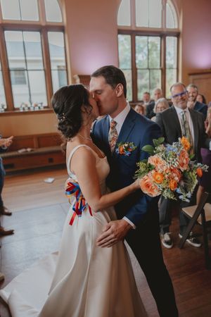 Romantic first kiss sequence of newlyweds at indoor wedding venue with warm lighting and colorful bouquet.