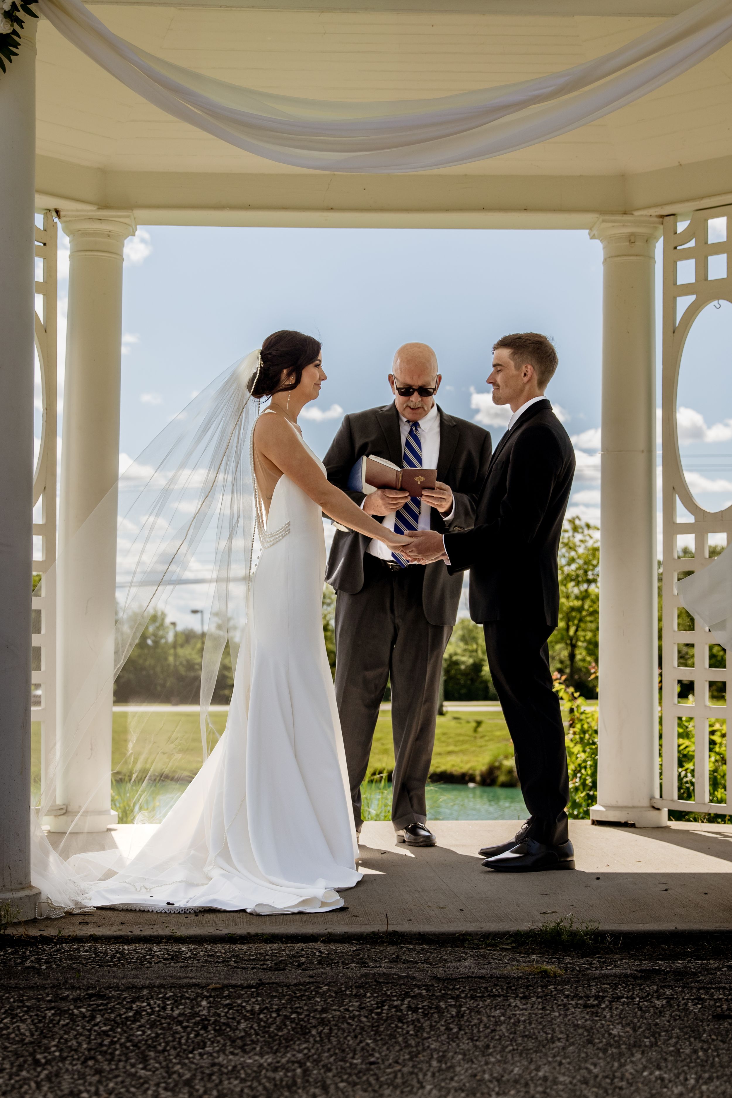 Wedding ceremony takes place under an elegant white gazebo with draped fabric and natural lighting.