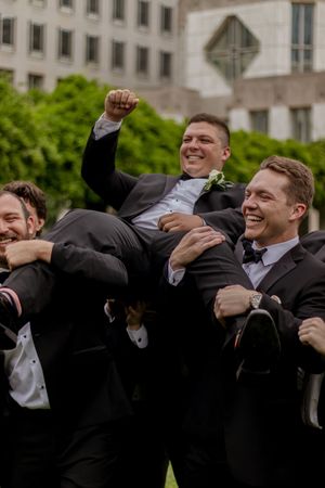 Groomsmen in black tuxedos celebrating and cheering joyfully outside a building.