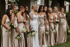 Bridesmaids in matching champagne colored dresses stand in a row holding white bouquets at outdoor wedding.