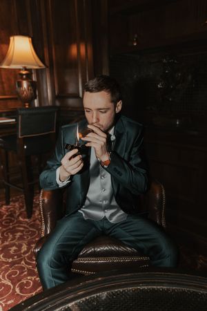 Moody portraits of groom in dark suit sitting in leather chair with dramatic lighting.