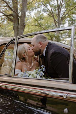 Sweet moment captured through vintage car window with white floral bouquet visible.