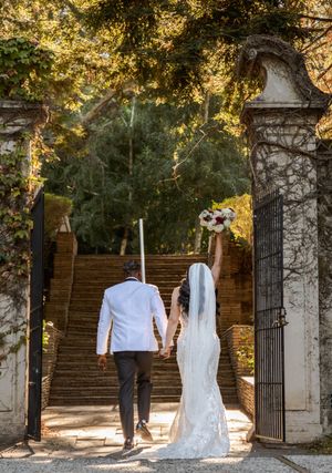Wedding dress and suit hanging between stone pillars and wrought iron gates in a garden setting.