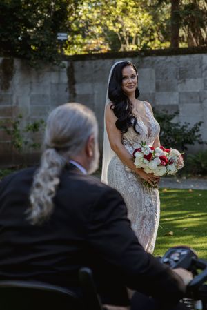 A bride holding a red and white bouquet smiles during an outdoor garden wedding ceremony.