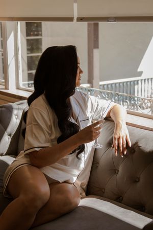 Person lounging on a grey tufted couch in a sunlit room with wine glasses visible.
