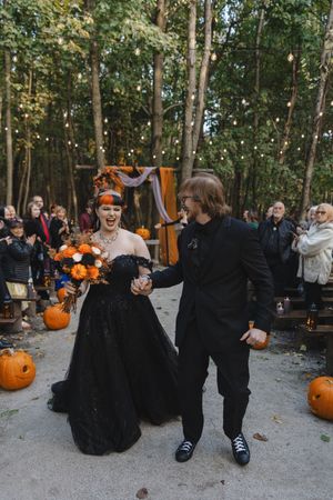A group wearing black costumes gathers outdoors with pumpkins during a fall celebration in a wooded area.