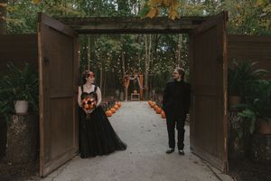 A couple walks down an outdoor wooden walkway decorated with orange flowers and lanterns for a wedding ceremony.