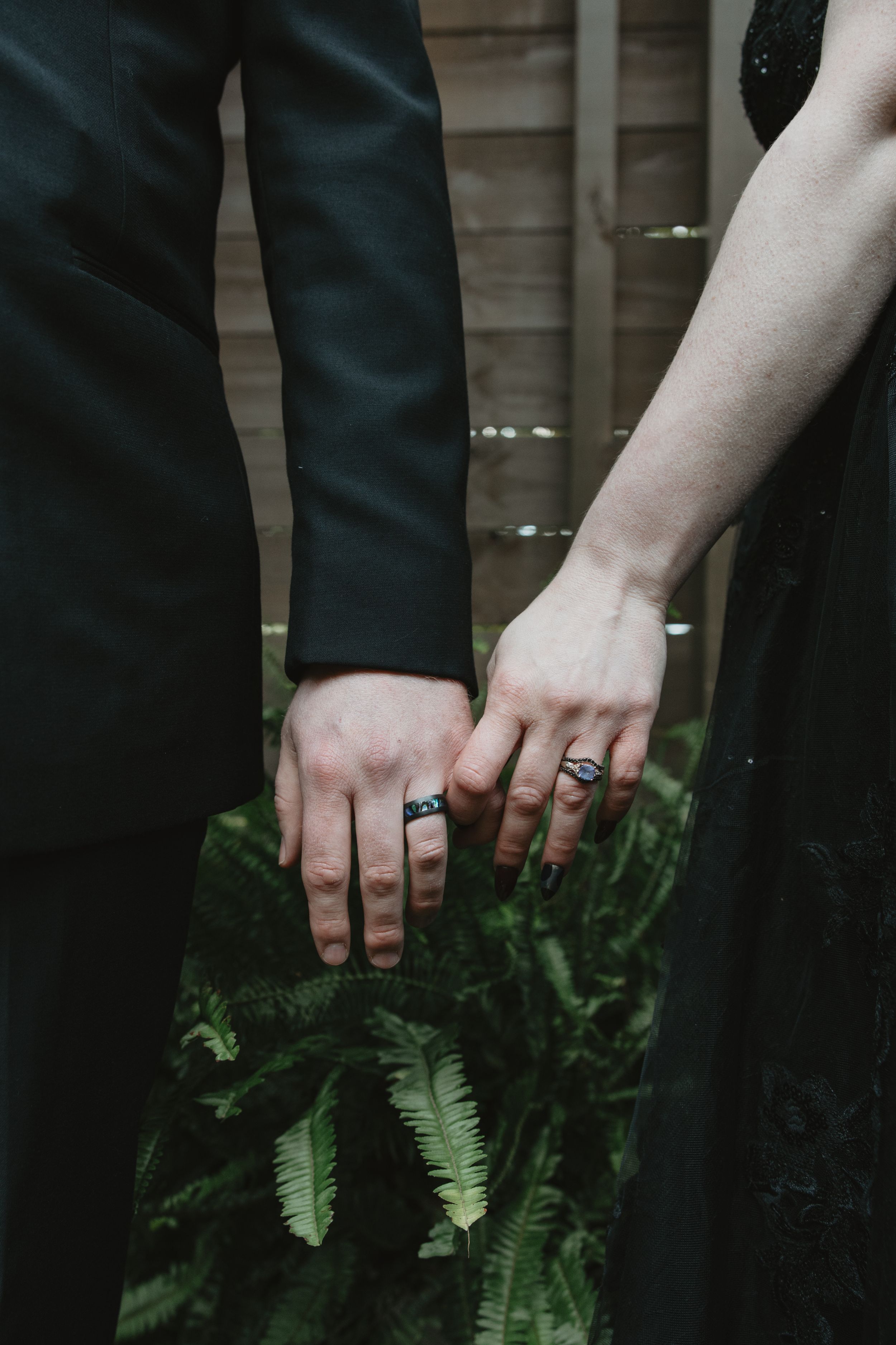 Close-up of hands wearing dark rings against black clothing and green fern leaves in a moody setting.