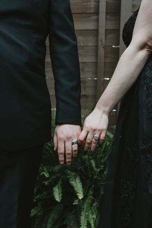 Close-up of two hands with wedding rings touching against a backdrop of green fern leaves.